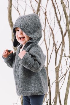 a young child standing in front of a tree wearing a gray sweater and blue pants