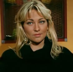 a woman with blonde hair and blue eyes looks at the camera while standing in front of a window