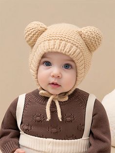 a baby wearing a knitted bear hat and sweater with suspenders, standing in front of a beige background