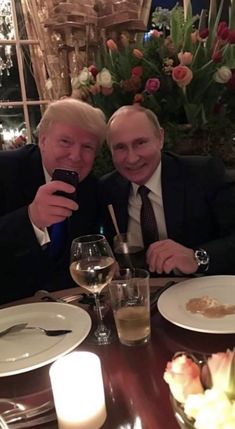 two men sitting at a dinner table taking a selfie with their cell phones in front of them