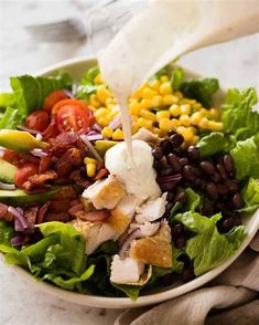 a salad with dressing being poured into it and garnished with bacon, lettuce, tomatoes, black beans, avocado, corn, cucumber wedges