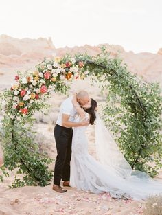 a bride and groom kissing under an arch decorated with flowers at their wedding in the desert