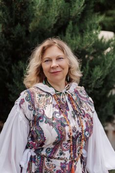 a woman standing in front of some trees wearing a white shirt with colorful patterns on it