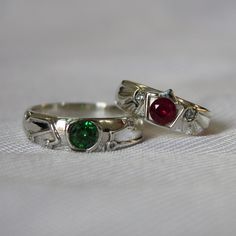 two silver rings with red and green stones on them sitting on a white cloth covered surface