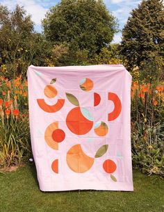 an orange and pink quilt on the grass in front of some flowers with trees behind it