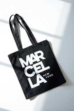 A canvas bucket tote bag in 100% European cotton, with a bottom panel for added capacity. The tote features an oversized MARCELLA NEW YORK graphic on one side, in white print on black canvas.[SPLIT] Our production team enjoys a living wage, a 40-hour work week with paid overtime, free health insurance and 20 days annual paid vacation. Black Rectangular Canvas Bag For Weekend, Casual Black Bags With Graphic Design, Black Cotton Tote Shoulder Bag, Large Capacity Black Cotton Canvas Bag, Cotton Tote Shoulder Bag With Logo Print, Black Canvas Weekend Bag, Black Canvas Bag For Weekend, Large Capacity Cotton Bucket Canvas Bag, Black Canvas Bag For The Weekend