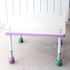 a white table with two purple and green legs on the top, sitting in front of a wall