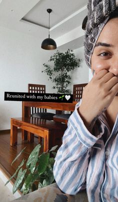 a woman covers her mouth while sitting at a table in front of a potted plant