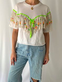 this fabulous reworked vintage festival shirt is so much fun! the white shirt is made of 100% cotton and features a crew neckline, half sleeves and a reworked raw cut cropped hem. there is fringed detailing throughout the front and the back of the shirt which features a bright neon green shoelace that ties in a bow on the centre front, real leather long fringes (around 5.5"), and colorful neon plastic beads to give it a boho festival flair. based on the tag, this shirt was presumably reworked/ha Spring Music Festival Top With Fringe, Spring Fringe Tops For Music Festival, Fringe Tops For Spring Music Festival, Summer Festival Green T-shirt, Spring Festival Tops With Beaded Fringe, Green Spring Tops With Tassels, Casual Fringe Crop Top For Festival, Spring Green Tops With Tassels, White Cropped Festival Top