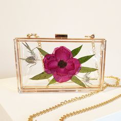 a pink flower is sitting in a clear box on a white surface with gold chains