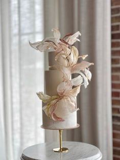 a three tiered wedding cake with pink flowers on the top and gold base, sitting on a table in front of a window