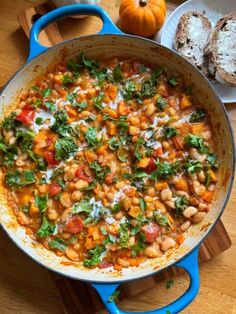 a blue pan filled with beans and vegetables on top of a wooden table next to bread