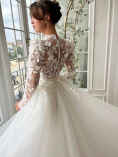 This bridal gown showcases a high neckline with a soft bow accent, paired with sheer long sleeves embellished with 3D floral appliqués. The fitted lace bodice is cinched at the waist with a delicate sash, flowing into a layered skirt featuring intricate lace beneath a tulle overlay. Ideal for weddings and formal occasions, this dress blends timeless sophistication with detailed floral artistry. Long Sleeve Lace Ball Gown With Lace Bodice, Long Sleeve Lace Gown With Illusion Neckline, Ceremony Gown With Long Lace Sleeves, Long Sleeve Dresses With Sheer Sleeves For Ceremony, Wedding Night Gown With Lace Bodice And Long Sleeves, Wedding Gown With Lace Sleeves, Long Sleeve Lace Bodice Gown For Wedding Night, Tulle Gown With Long Lace Sleeves, Long Sleeve Gown With Lace Bodice For Wedding Night