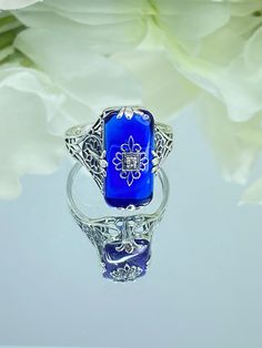 Sapphire Blue Glass Sterling Silver Ring Grace Design#D233 This is a lovely antique Edwardian era inspired filigree ring. This gorgeous ring is created in stunning sapphire blue glass. The glass is embellished with a delicate floral filigree. The top of the ring holds a center set 1mm round cut gemstone (Choose from white Cubic Zirconia, lab created white Moissanite, or Natural/Genuine White Diamond). The blue glass is 18mm x 19mm. The ring sits 18mm north-south and 10mm east-west on the finger. The inside of the band is marked 925 for sterling silver. The band's intricate open work filigree has hints of floral influence and geometric design running its entire length. The curves and swirls accent the beautiful smooth stone. The center filigree highlights the sparkling gemstone. This ring r Blue Victorian Filigree Jewelry, Antique Blue Rings With Rose Cut Diamonds, Sapphire Filigree Ring For Formal Occasions, Vintage Blue Filigree Ring For Anniversary, Antique Blue Rings For Wedding, Formal Sapphire Filigree Ring With Intricate Design, Victorian Blue Rings With Rose Cut Diamonds, Vintage Blue Sapphire Ring With Intricate Design, Ornate Filigree Ring With Rose Cut Diamonds For Gift