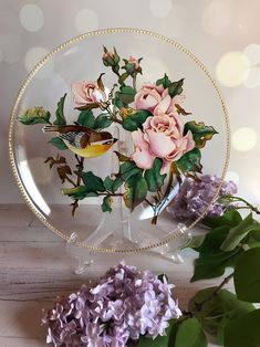 a glass plate with flowers on it next to some green leaves and purple lilacs