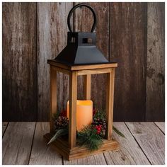 a wooden lantern with a lit candle inside on a wood table next to pine cones and greenery