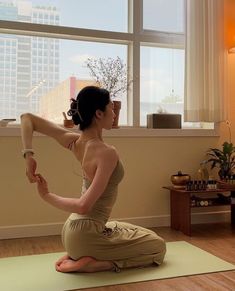 a woman sitting on a yoga mat in front of a window with her arms stretched out