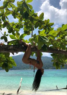 a woman hanging upside down from a tree branch on the beach with blue water in the background