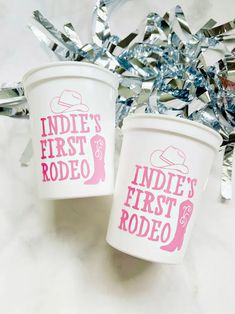 two white cups with pink designs on them sitting next to some tinsel and confetti