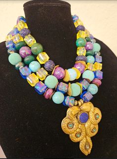 Lightweight with stunning colors, this triple strand statement necklace is destined to become one of your favorites! The featured colors are blue, yellow, purple, and green. The components include lucite cubes, magnesite, emerald, etched sapphire, mint green designer beads, white moonstone and copper and gold tone wavy metal spacers. The pendant is a hand crafted Tibetan brass repousse double dragon with inlaid lapis lazuli and turquoise. Adjusts from 17-20" and I can add additional chain links if requested. Closes with a gold tone lobster claw clasp and a 3" extender chain. From the Atelier of Kat Kouture! A perfect size so as not to overwhelm a petite woman. Colorful Beads Multi-strand Bib Necklaces For Jewelry Making, Colorful Beads Multi-strand Bib Necklace For Jewelry Making, Unique Multi-strand Necklace With Variations, Spiritual Multi-strand Jewelry With Large Beads, Handmade Double Strand Beaded Necklaces, Handmade Double Strand Beaded Necklace, Unique Multicolor Double Strand Necklace, Vibrant Blue Round Bead Jewelry, Spiritual Multicolor Jewelry With Unique Variations