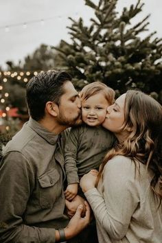 a man and woman kiss their son as they sit in front of a christmas tree