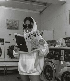 a woman in a hoodie reading a newspaper while standing next to washers and dryer machines