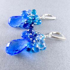 "Stunning cobalt blue lab created topaz dangle below a generous cluster of sparkling briolettes consisting of turquoise blue, navy blue, peacock green and sky blue quartz. I have used all sterling silver to keep them fresh. The gems are topped off with another blue topaz gem and a sterling silver bead. The blue topaz measure 20x11mm. The total earring length is 2 1/8\" (54mm.) from the top of the lever back ear wires." Blue Gemstone Cluster Earrings As Gift, Blue Gemstone Cluster Earrings For Gift, Blue Briolette Earrings For Wedding, Blue Gemstone Accented Drop Earrings, Blue Briolette Gemstone Earrings, Blue Drop Earrings With Gemstone Accents, Blue Peacock, Diy Making, Making Earrings