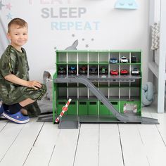 a young boy sitting on the floor next to a green shelf with toy cars in it