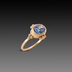 This stunning ring features a vibrant oval shaped blue sapphire, framed by twelve sparkling white diamonds. This very special gem is set in 18k yellow gold on a 1.5mm band. The center stone measures 7mm x 9mm, and weighs 3.59 ct. Matte finish. Yellow Gold Sapphire Ring With Diamond Oval Cabochon, Yellow Gold Sapphire Diamond Ring With Oval Cabochon, Oval Cabochon Sapphire Ring In Yellow Gold With Diamond, 14k Gold Sapphire Ring With Rose Cut Diamonds, Modern Sapphire Ring With Rose Cut Diamonds, Tanzanite Yellow Gold Rings With Diamond Accents, Oval Sapphire Diamond Ring With Bezel Setting, Yellow Gold Tanzanite Rings With Diamond Accents, Oval Yellow Gold Sapphire Ring With Single Cut Diamonds