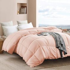 a bed with pink comforter and pillows in a room next to a window overlooking the ocean