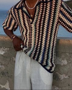 a man standing next to the ocean wearing a crocheted shirt and white pants