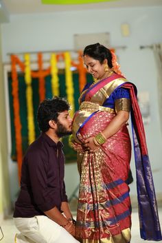 a man kneeling down next to a woman in a red and blue sari with her hand on her belly