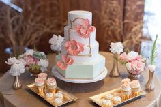 a wedding cake with pink flowers and gold trimmings on a table next to cupcakes