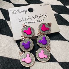 three pairs of pink and purple mickey mouse earrings on top of a black and white checkered tablecloth