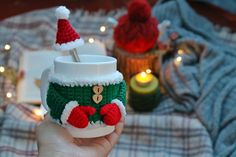 a hand holding a cup with a knitted santa hat on it and two candles in the background