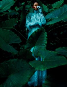 a woman in a blue dress is standing among green plants and leaves with her phone to her ear