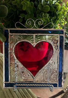 a red heart shaped glass hanging from a metal frame with green plants in the background