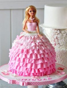 a barbie doll sitting on top of a pink and white tiered dress next to a wedding cake