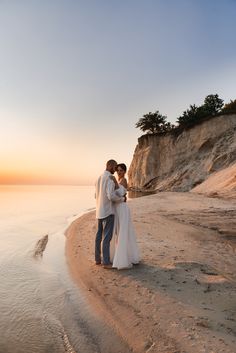 a bride and groom standing on the beach in front of an ocean cliff at sunset