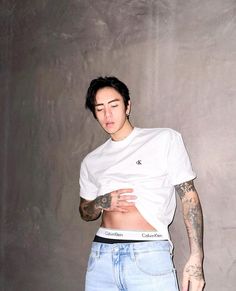 a young man with tattoos standing in front of a wall wearing blue jeans and a white t - shirt