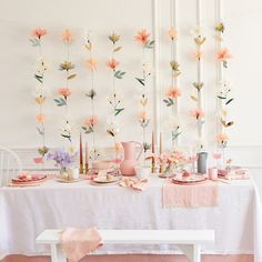 a white table topped with pink and yellow flowers next to a wall covered in paper flowers