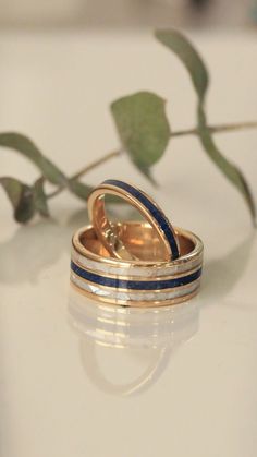 A custom wedding ring set from Staghead Designs sits on a white tile background. This 14K yellow gold set features our "Rio" design wedding band with a centered lapis lazuli inlay & two outer mother of pearl inlays. The other ring is our narrow "Eterna" band featuring a lapis lazuli inlay. This fully customizable set can also be made in 14K white or rose gold & with a wide variety of unique inlay materials including wood, antler, turquoise, or even materials you send in! Whiskey Barrel Wedding Ring, Antler Wedding Rings, Matching Wedding Ring Sets, Matching Wedding Ring, Handcrafted Engagement Ring, Wood Wedding Ring, Mens Gold Wedding Band, Matching Wedding Rings, Staghead Designs