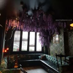 a living room filled with lots of furniture and purple flowers hanging from the ceiling above