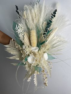 an arrangement of white flowers and feathers on a gray background with someone's hand holding it