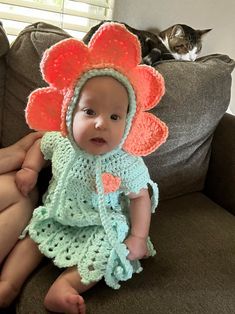 a baby wearing a crocheted flower hat sitting on a couch next to a cat