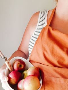 a woman in an orange dress holding a white bowl filled with red and yellow apples