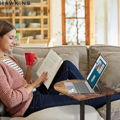 a woman sitting on a couch holding a cup and looking at a book while using a laptop computer