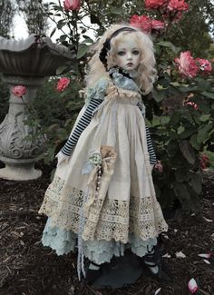 a doll is standing in front of some flowers and bushes, wearing a dress with laces on it