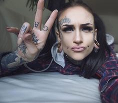 a woman with tattoos and piercings on her face making the v sign while laying in bed