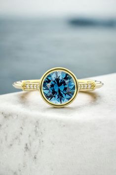 a blue diamond ring sitting on top of a white marble slab with water in the background